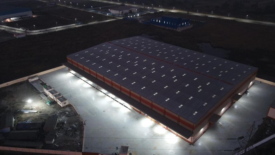 The image shows an aerial view of a large warehouse complex at dusk or nighttime. The warehouse building has a rectangular structure with a light gray roof dotted with evenly spaced skylights or vents. The surrounding area includes paved grounds with stree