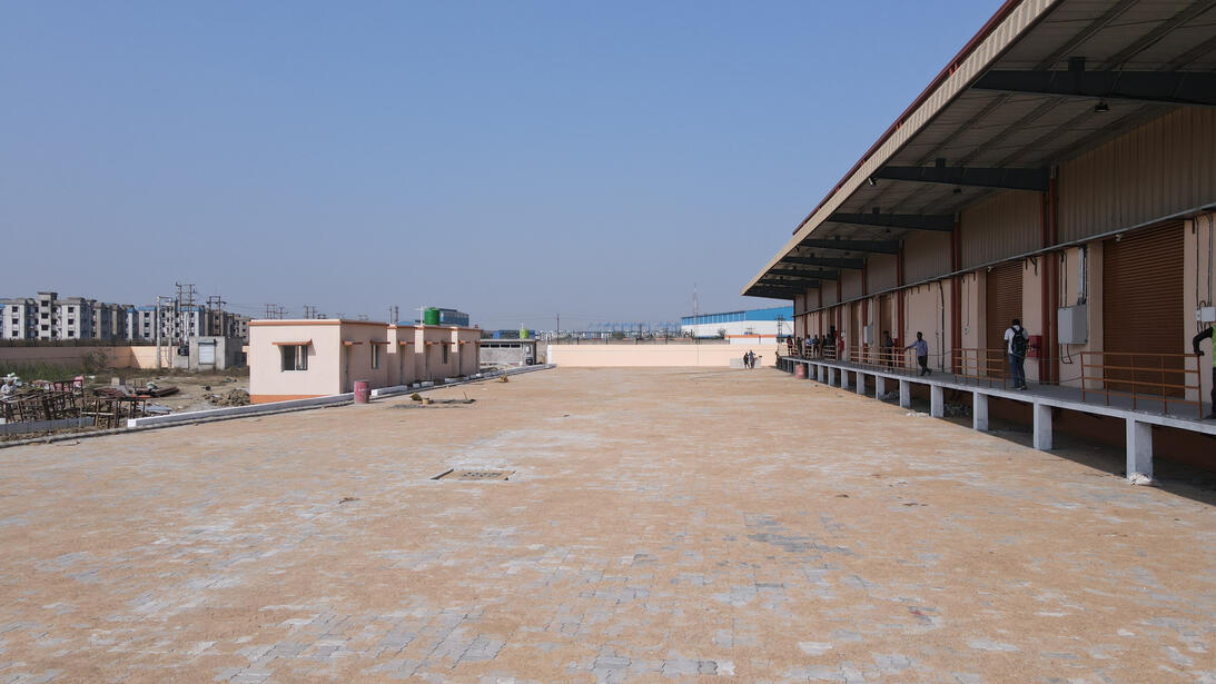 The image shows a daytime view of an industrial warehouse facility. On the right side, a large warehouse building with a long row of loading bays can be seen. Several people are standing near the loading area, which is elevated with a platform and sheltere