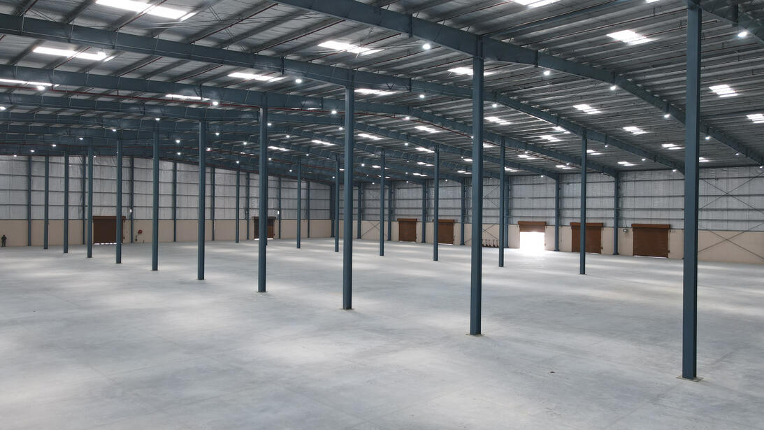 The image shows the interior of a large, empty warehouse with high ceilings and a spacious open floor plan. The warehouse features metal columns and beams, with a roof supported by a grid of steel framing. Several ceiling lights and skylights provide ample